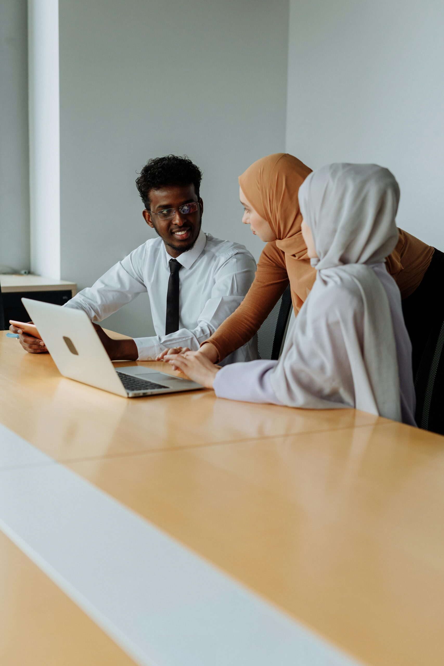 Three colleagues in an office discussing work, including two women in hijabs and a man with a laptop.